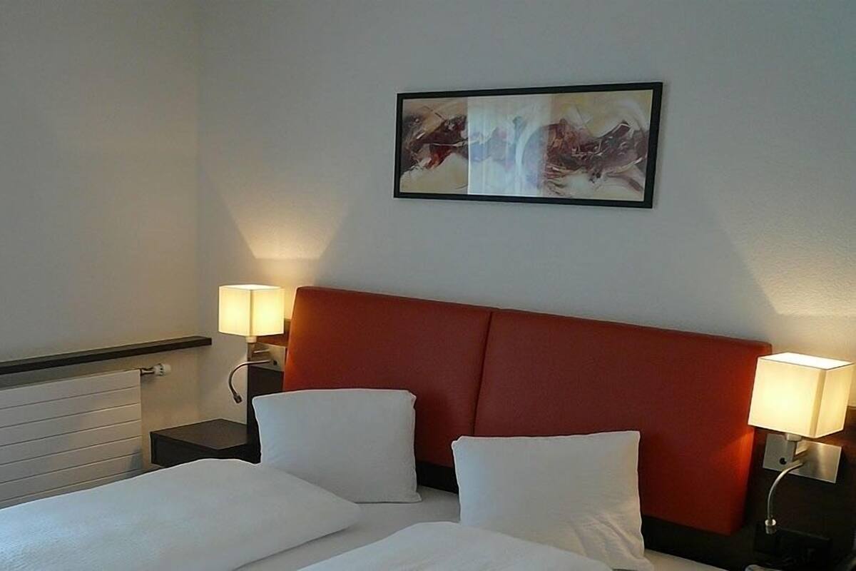 A hotel room with a bed, two bedside tables with lamps, a red headboard, and a framed abstract painting on the wall.