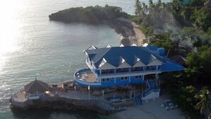A large, multi-story building with a blue roof, situated on a beach with clear water and a rocky outcrop nearby. - Blue Corals Beach Resort (Daanbantayan)