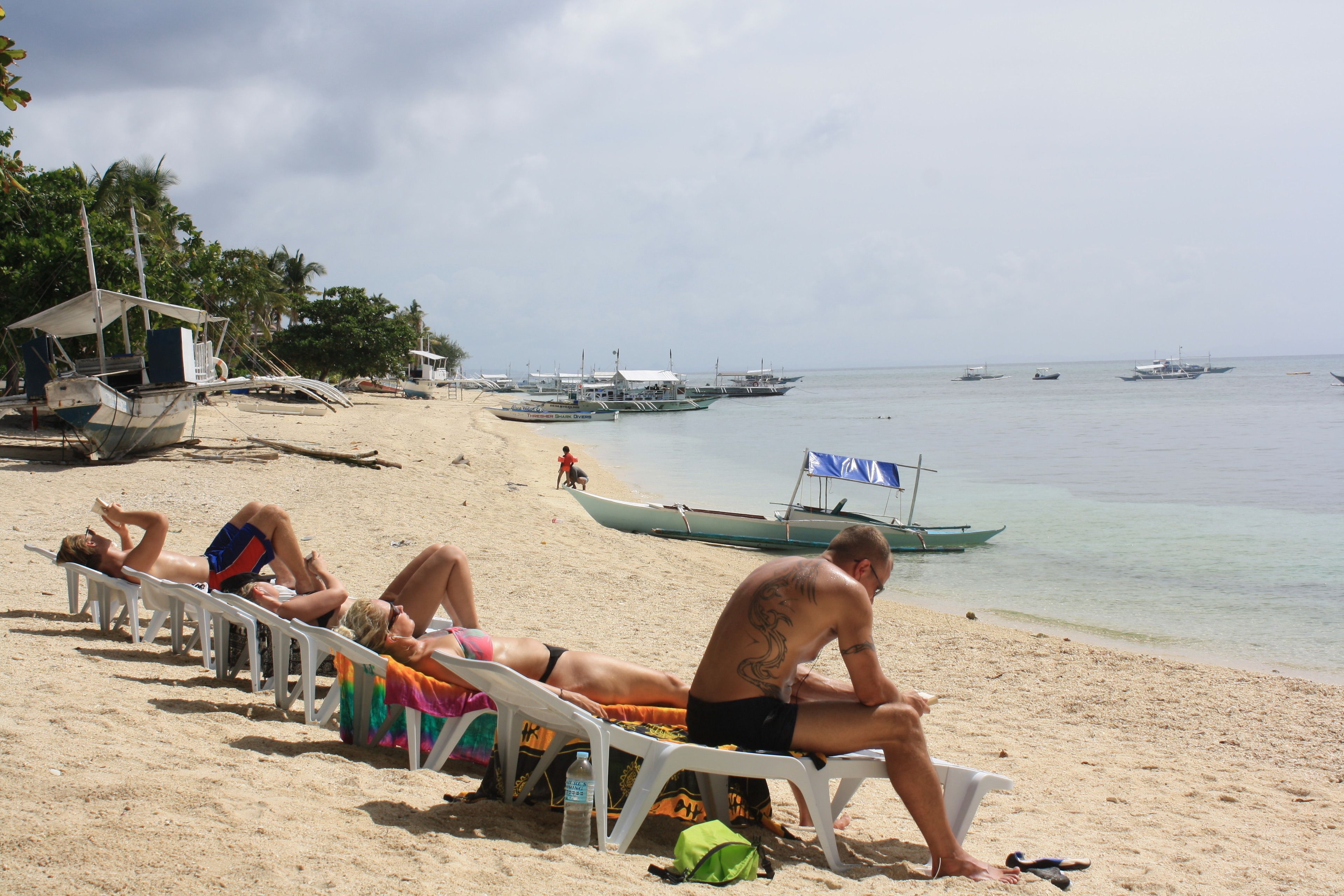 A beach with people sunbathing on lounge chairs, a small hut, and several boats in the water.