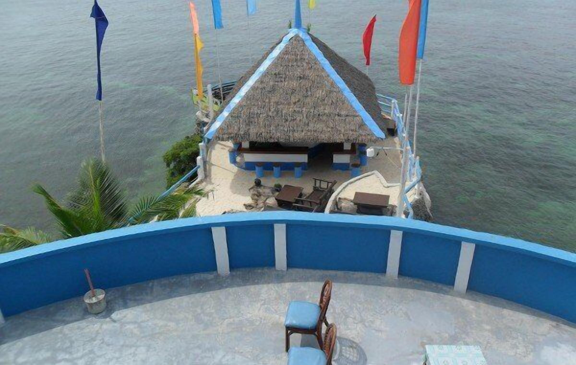 A thatched-roof hut with a blue and white exterior, surrounded by a pool area with lounge chairs, set against a backdrop of calm water and colorful flags.