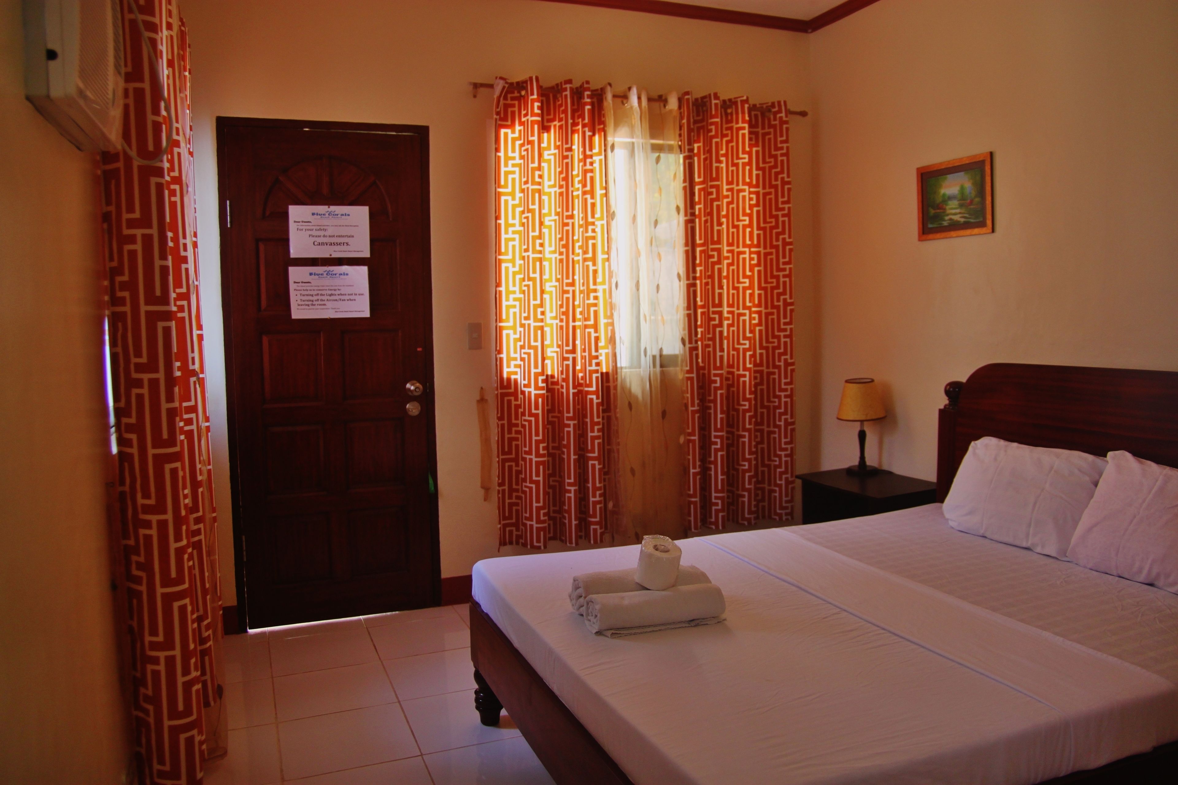 A hotel room with a bed, a wooden headboard, a nightstand with a lamp, and a door with a sign.
