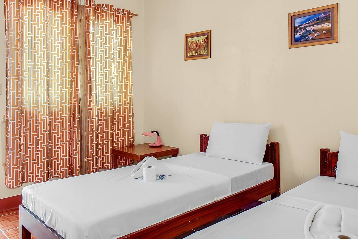 Habitación de hotel con dos camas individuales, un cabecero de madera, una mesita de noche con una lámpara rosa y un cuadro en la pared.