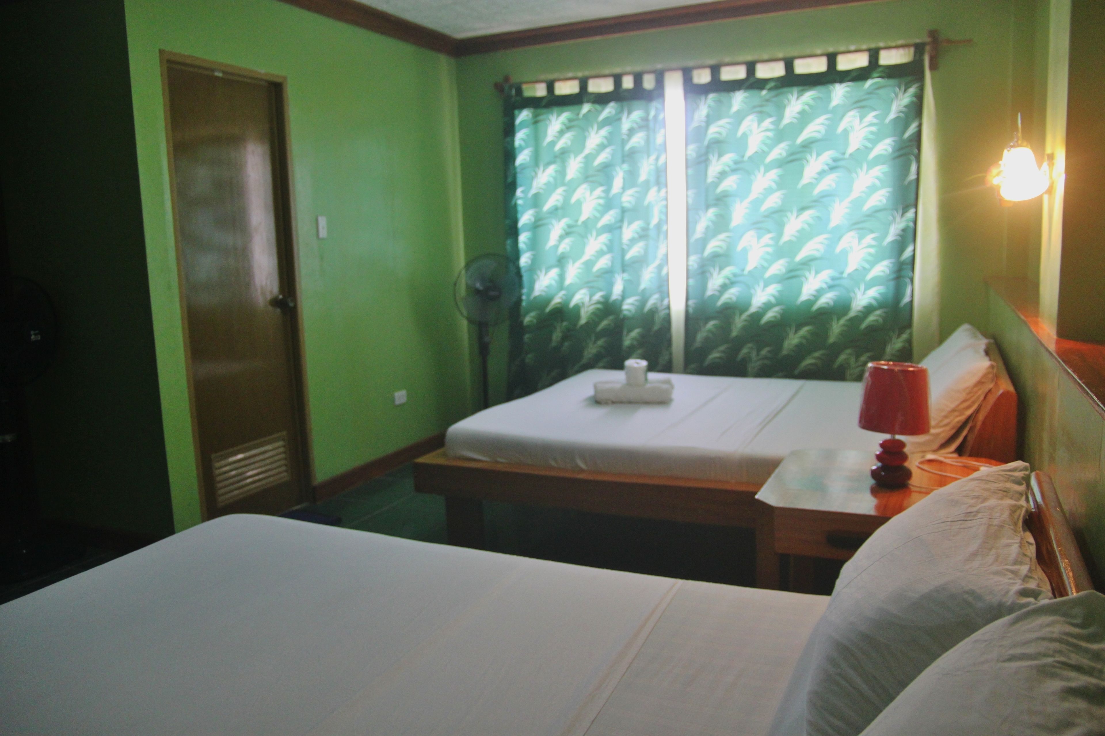 Una habitación con una cama, una mesita de noche con lámpara, un ventilador y una ventana con cortinas estampadas.