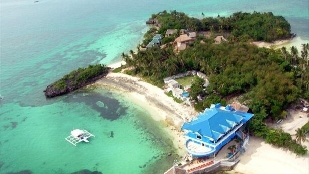 An aerial view of a tropical island with a beach, clear turquoise waters, and a small boat. - Blue Corals Beach Resort (Daanbantayan)