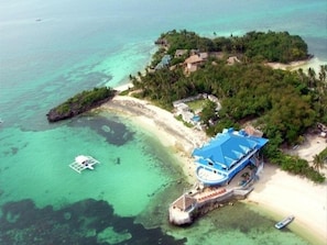An aerial view of a tropical island with a beach, clear turquoise waters, and a small boat. - Blue Corals Beach Resort (Daanbantayan)