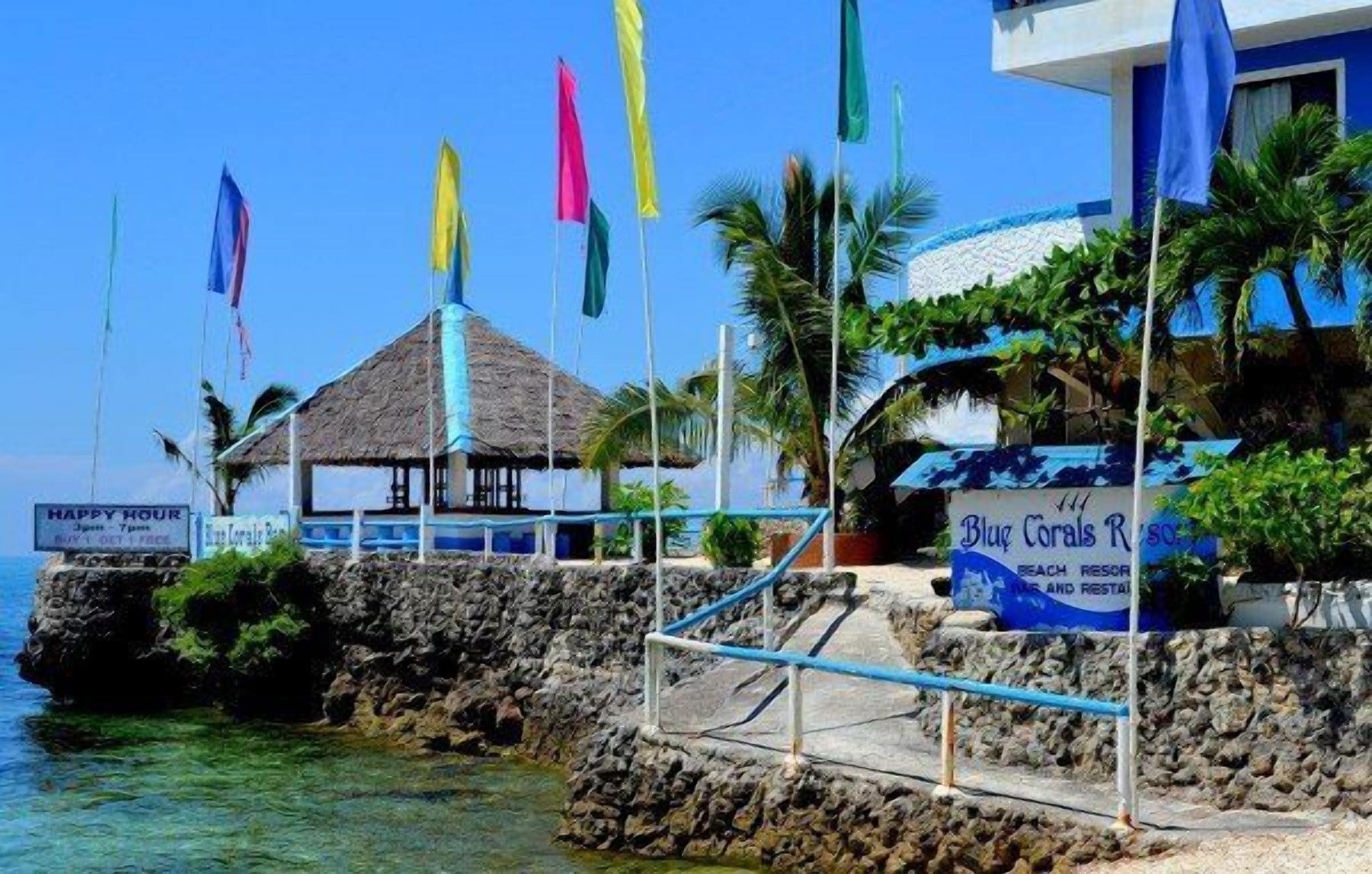 A beachfront property with a thatched-roof structure, a signboard for 'Blue Corals Resort', and multiple colorful flags.