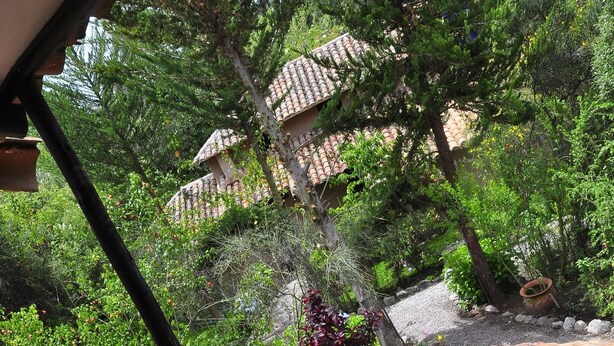 A wooden deck with a cushioned chair and a small table, overlooking a garden with trees and a building with tiled roofs. - Kuychi Rumi (Urubamba)