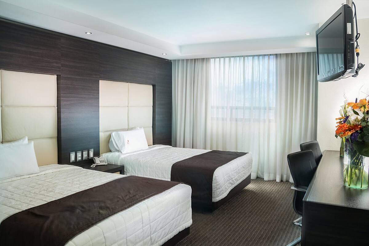 Ein Hotelzimmer mit zwei Betten, einem Schreibtisch, einem Stuhl, einem Fernseher und einem Fenster mit Vorhängen.