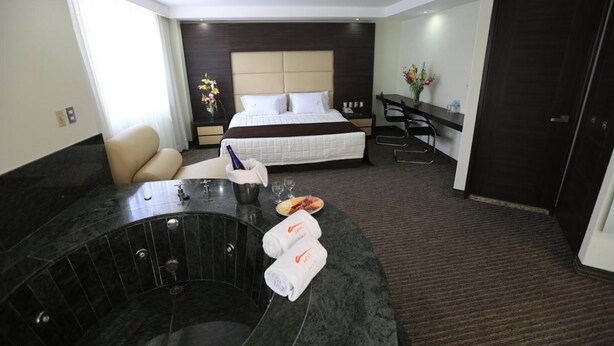 A hotel room with a bed, a sitting area with a chair, a small table with a bottle and glasses, a hot tub, and a door.