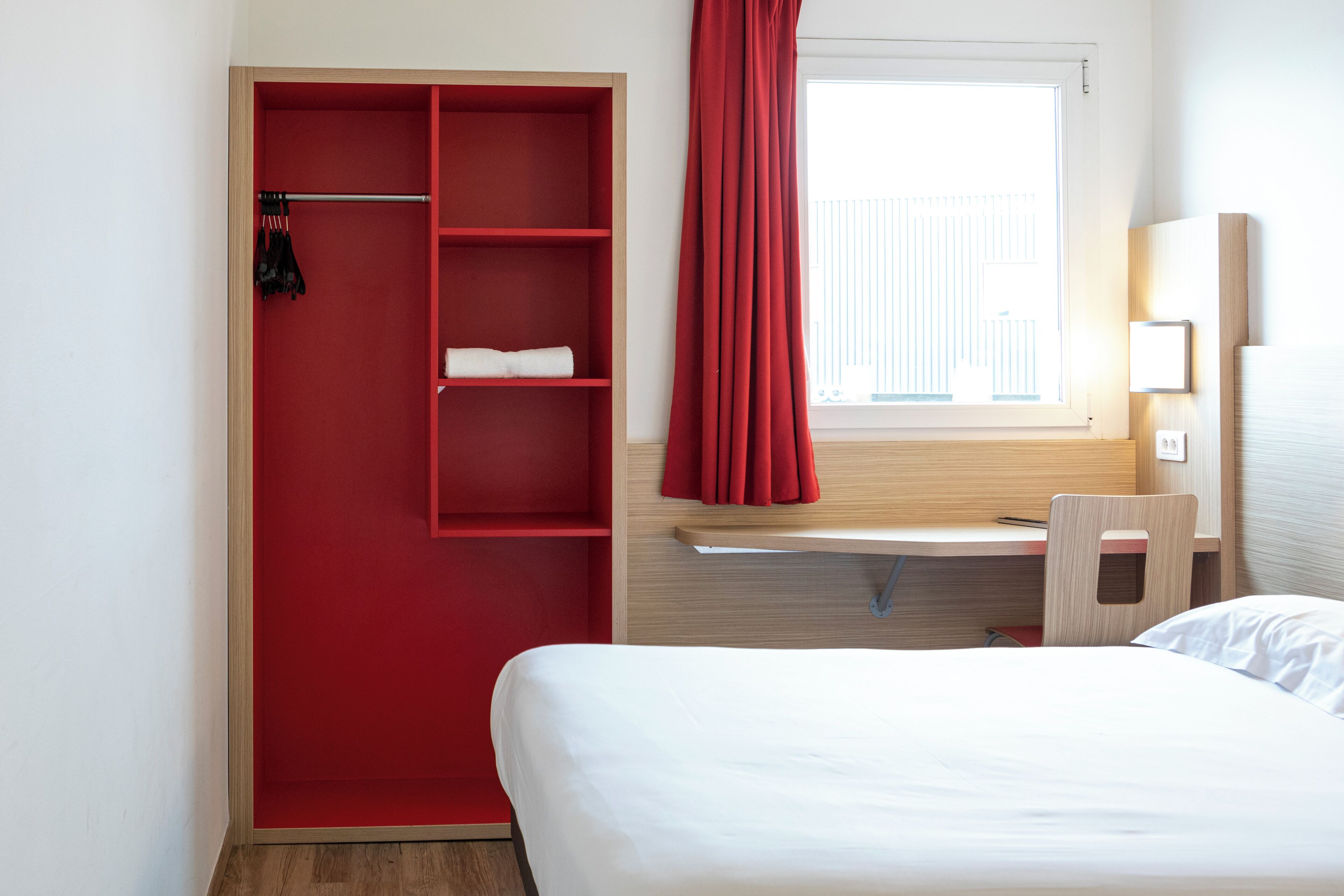A hotel room with a bed, a desk with a chair, a wardrobe with shelves, and a window with curtains.