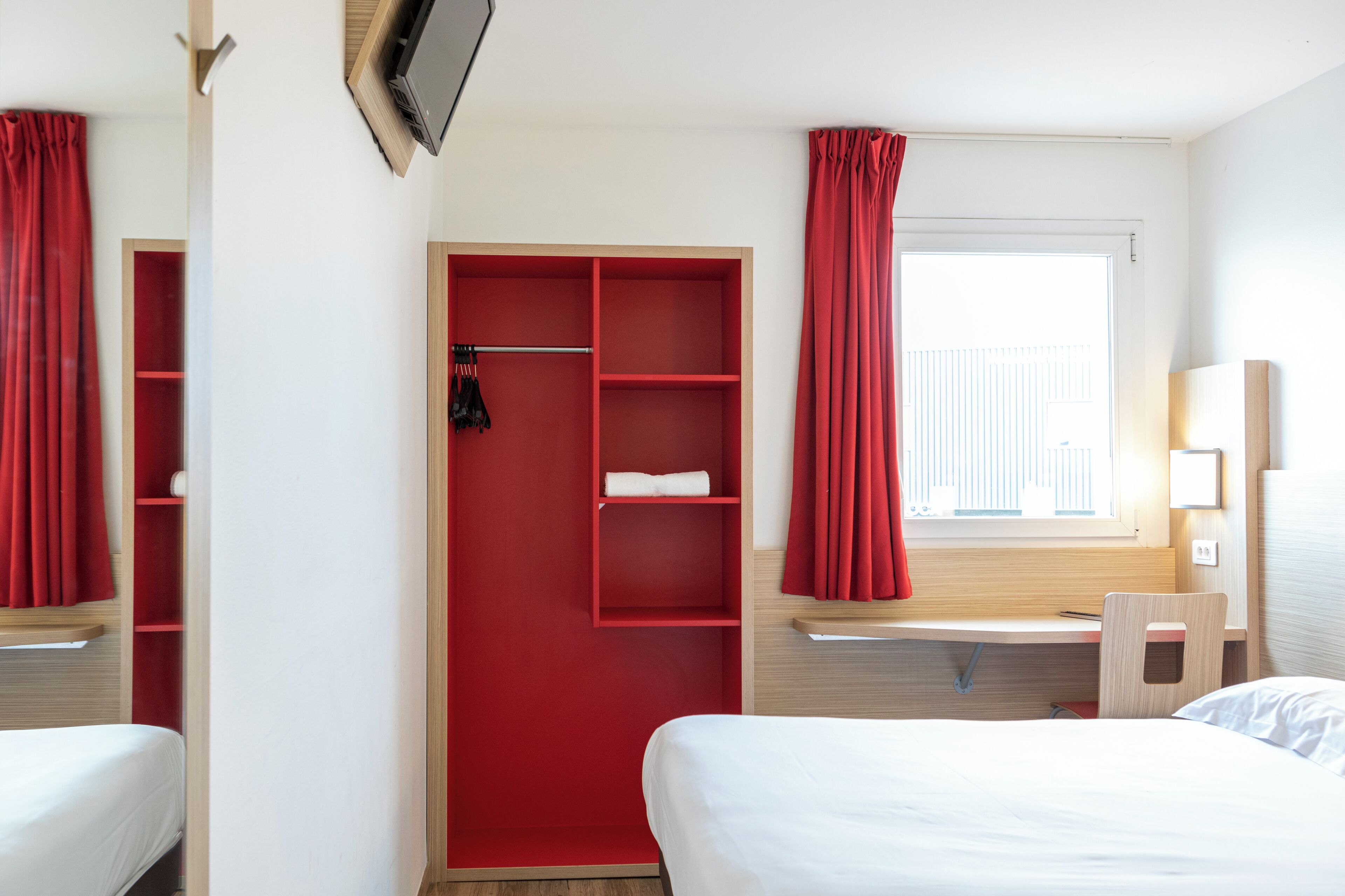 A hotel room with a bed, a desk, a chair, a TV, a wardrobe, and a window with curtains.
