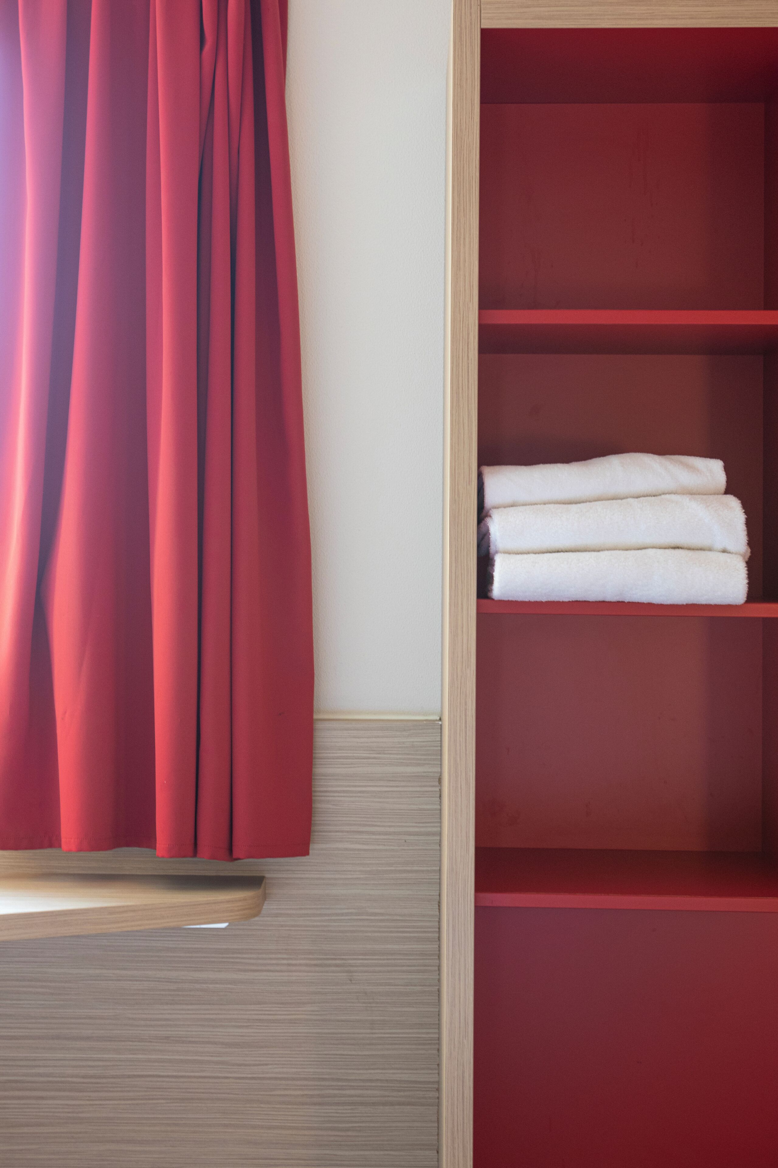 A room with a red curtain, a wooden shelf with folded white towels, and a red shelf unit.