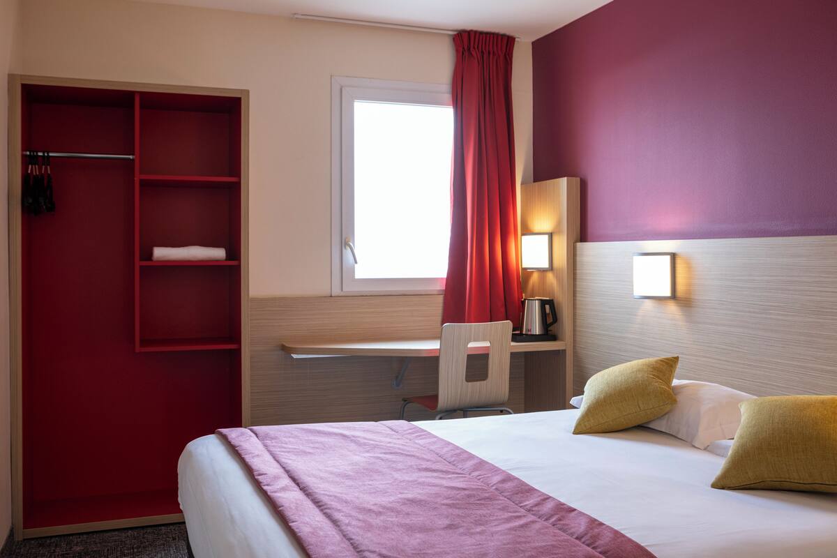 Une chambre d’hôtel comprenant un lit, un bureau, une chaise, une armoire et une fenêtre avec des rideaux.