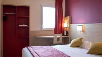 Quarto de hotel com cama, escrivaninha, cadeira, guarda-roupa e janela com cortinas.