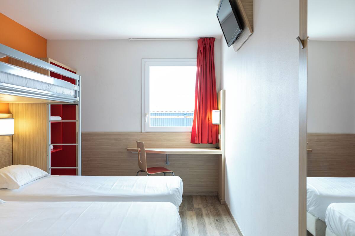 Une chambre d’hôtel avec des lits superposés, un bureau, une chaise, une télévision et une fenêtre avec des rideaux.