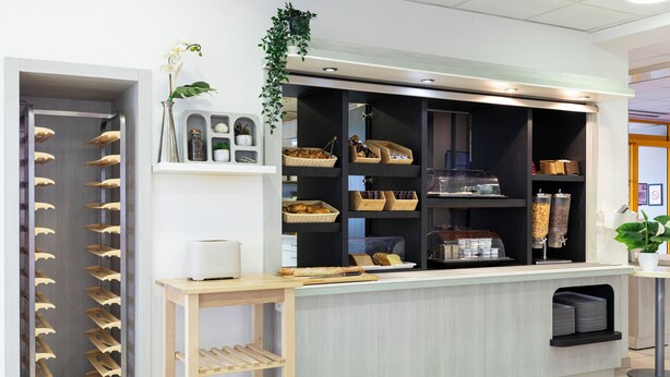 A reception area with a counter, shelves with baskets, a plant, and a rack of trays. - The Originals ACCESS Tendance Hôtel (Andrezieux-Boutheon)