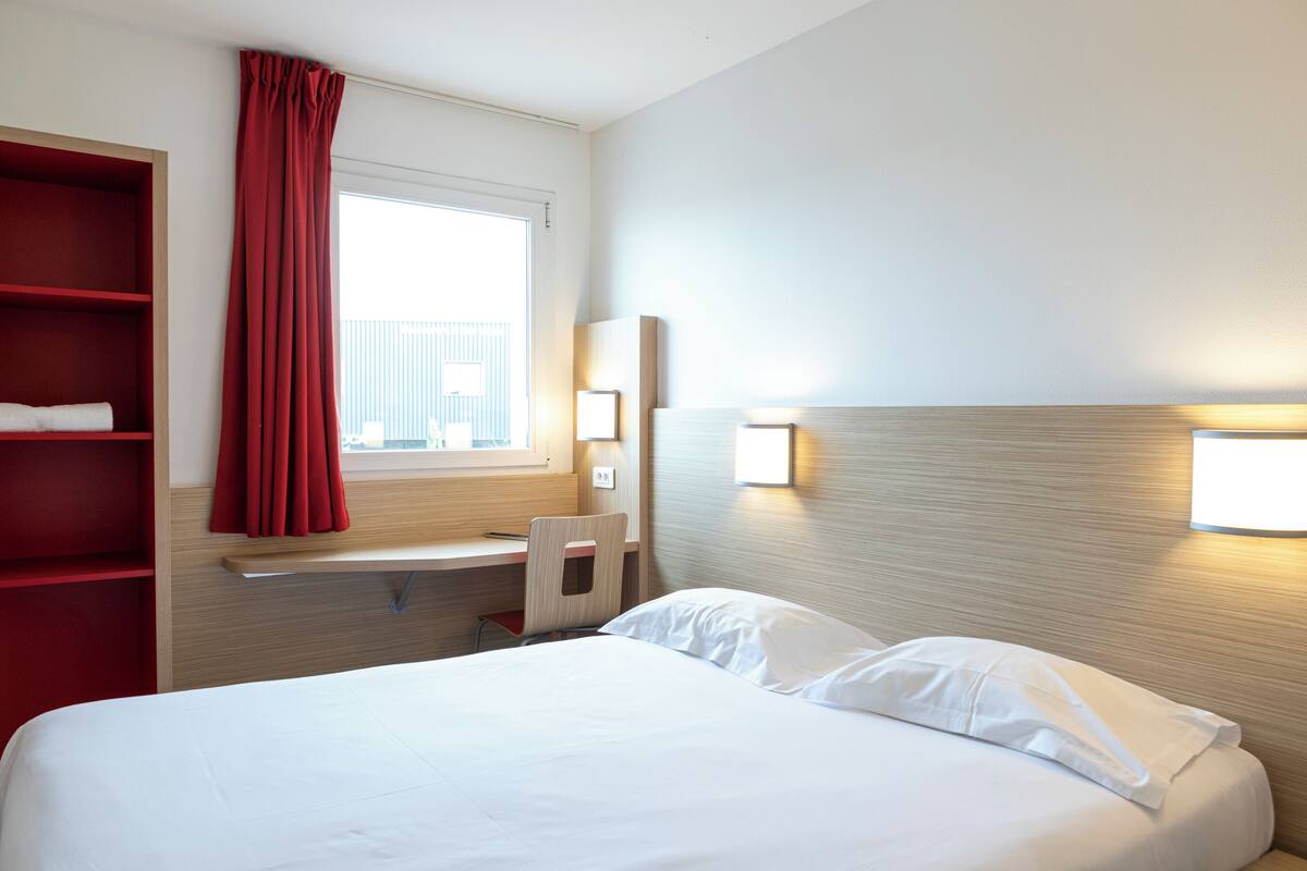 Une chambre d’hôtel avec un lit, un bureau, une chaise, une étagère rouge, une fenêtre avec des rideaux et des lampes fixées au mur.