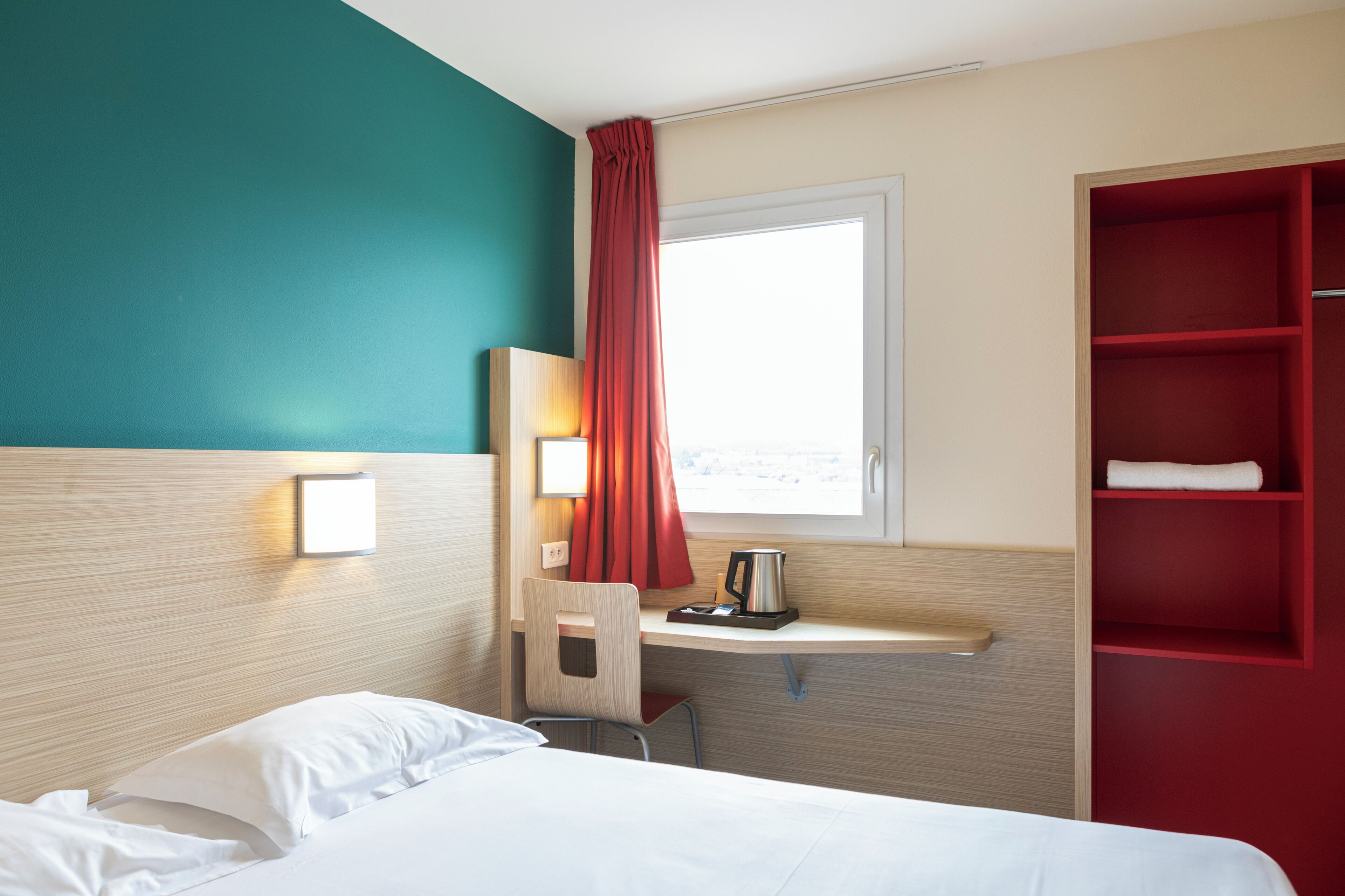 A hotel room with a bed, a desk with a chair, a red shelf, a window with curtains, and a wall-mounted light.