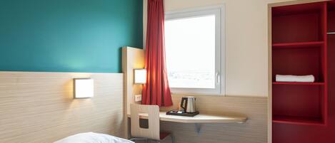 Habitación de hotel con cama, escritorio con silla, estante rojo, ventana con cortinas y luz fijada a la pared.