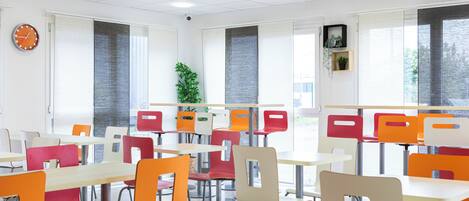 Una cafetería con mesas y sillas, un reloj y ventanas con persianas.