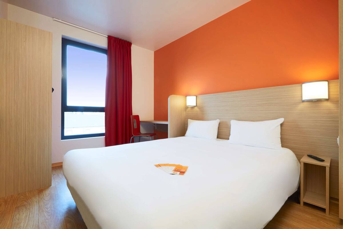 Quarto de hotel com uma cama grande, uma escrivaninha com cadeira, uma janela com cortinas vermelhas e um ponto de luz fixado na parede.