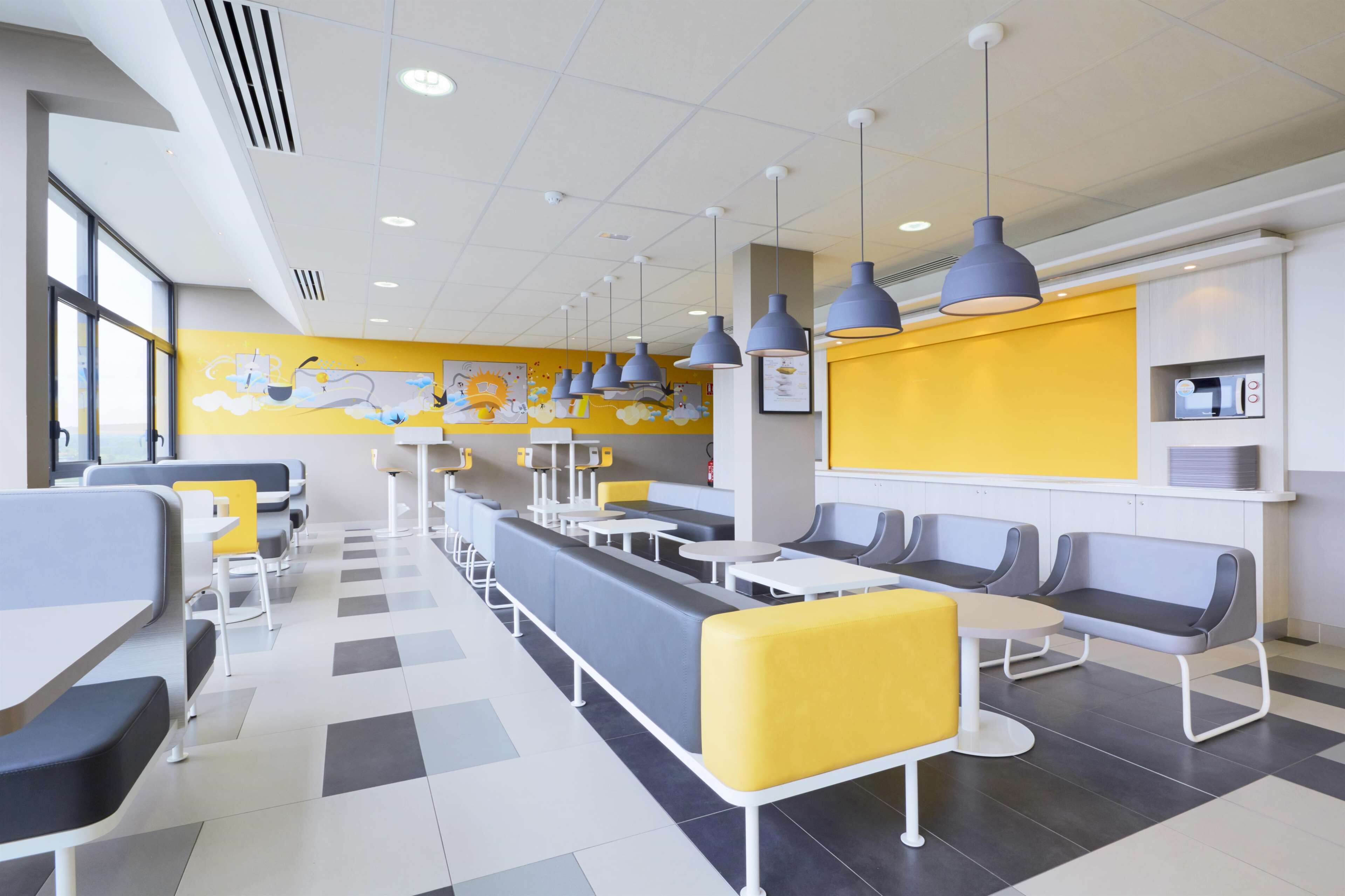 A modern cafeteria with long communal tables and booths, a yellow accent wall, and overhead pendant lighting.