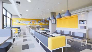 Ett modernt kafeteriaområde med långa, gemensamma bord och avskilda sittplatser, en gul accentvägg och hängande lampor i taket.