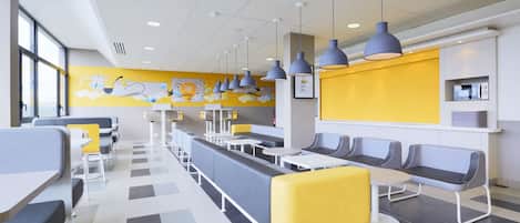 A modern cafeteria with long communal tables and booths, a yellow accent wall, and overhead pendant lighting.