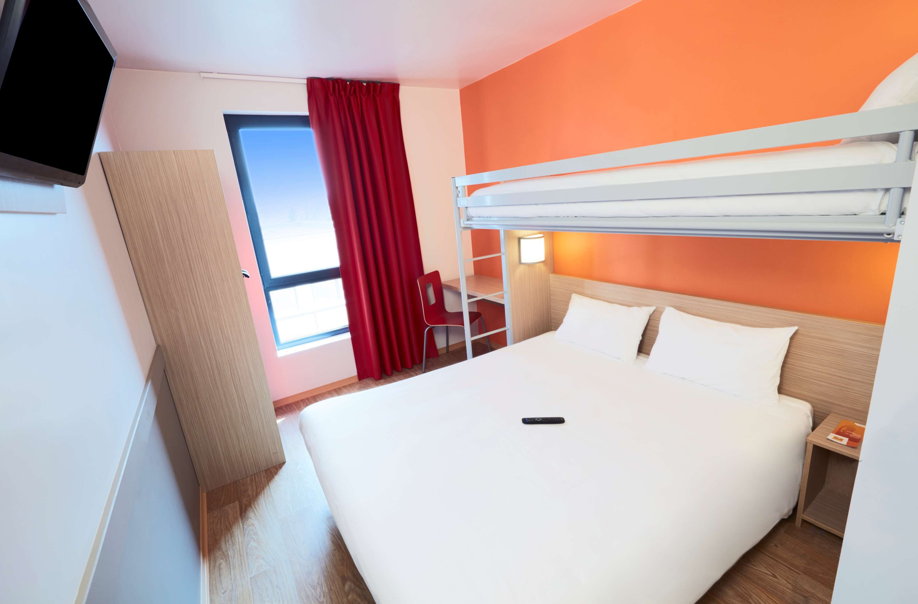 A hotel room with a bunk bed, a desk with a chair, a TV, and a window with red curtains.