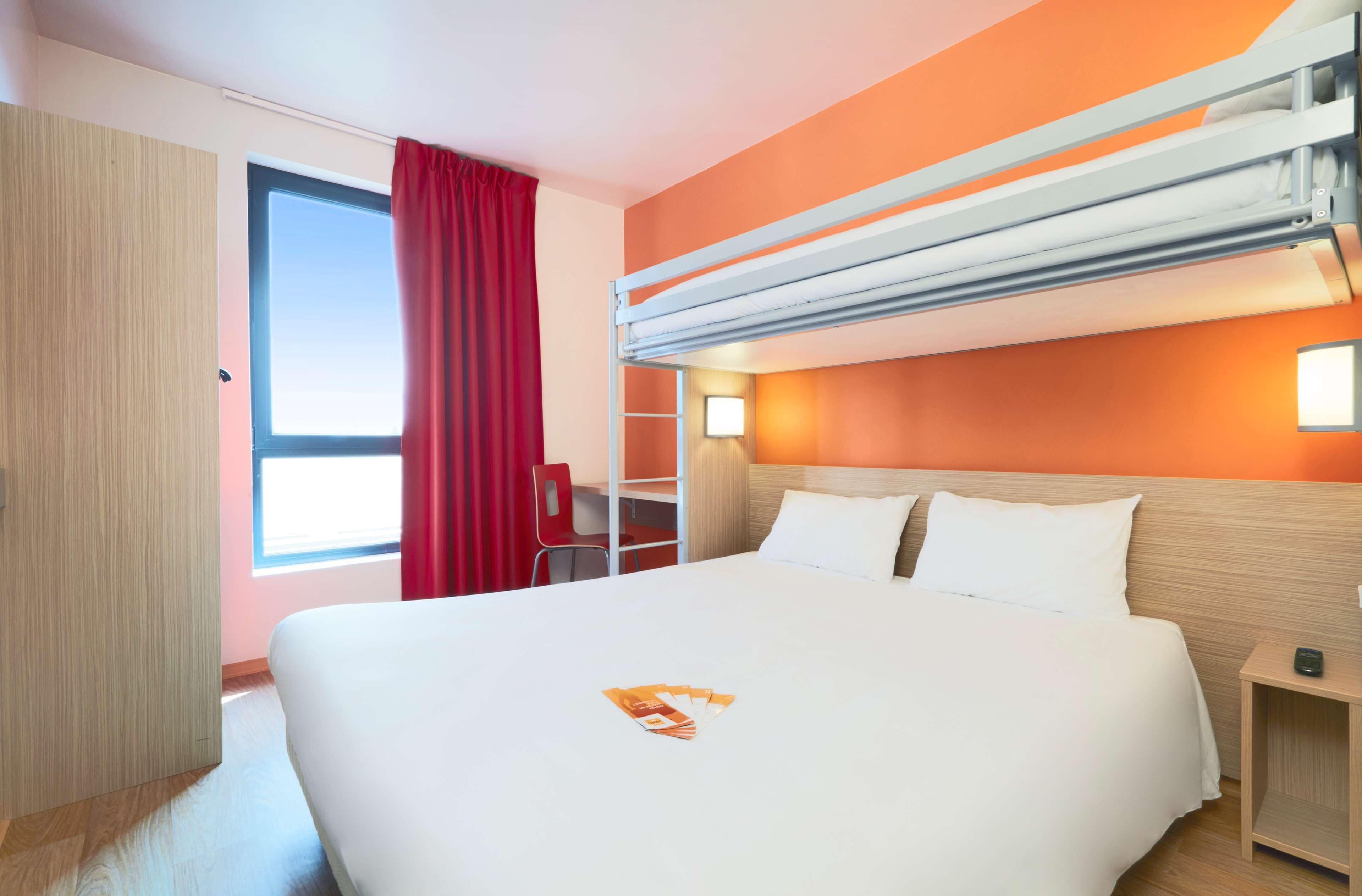 A hotel room with a bed, a desk, a chair, and a window with red curtains.