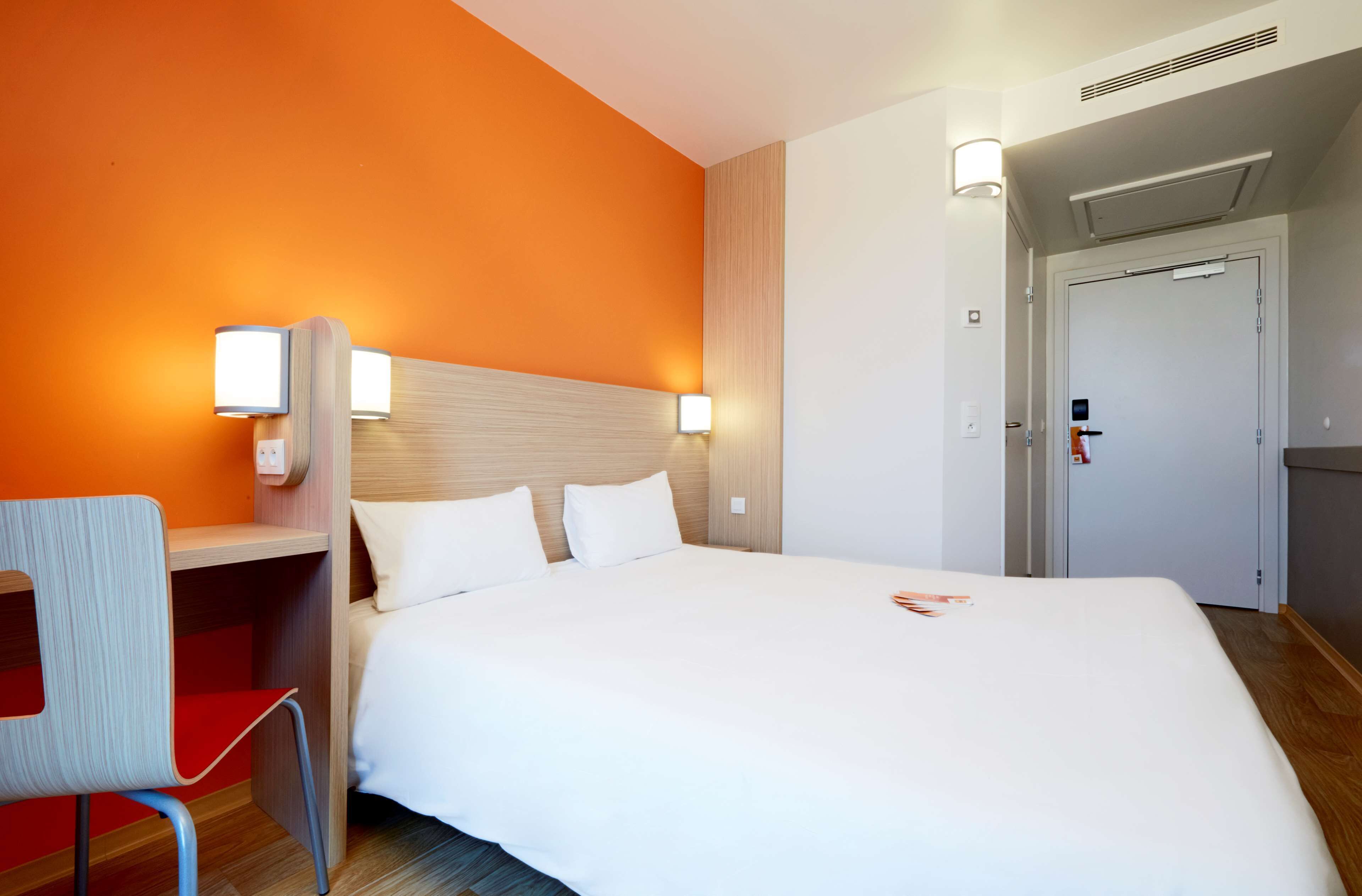 A hotel room with a large bed, a desk with a chair, and a door.