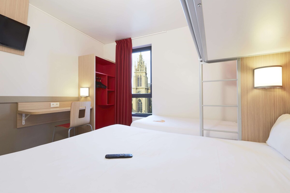 A hotel room with a bed, a desk, a chair, a TV, a window with a view of a church, and a red curtain.