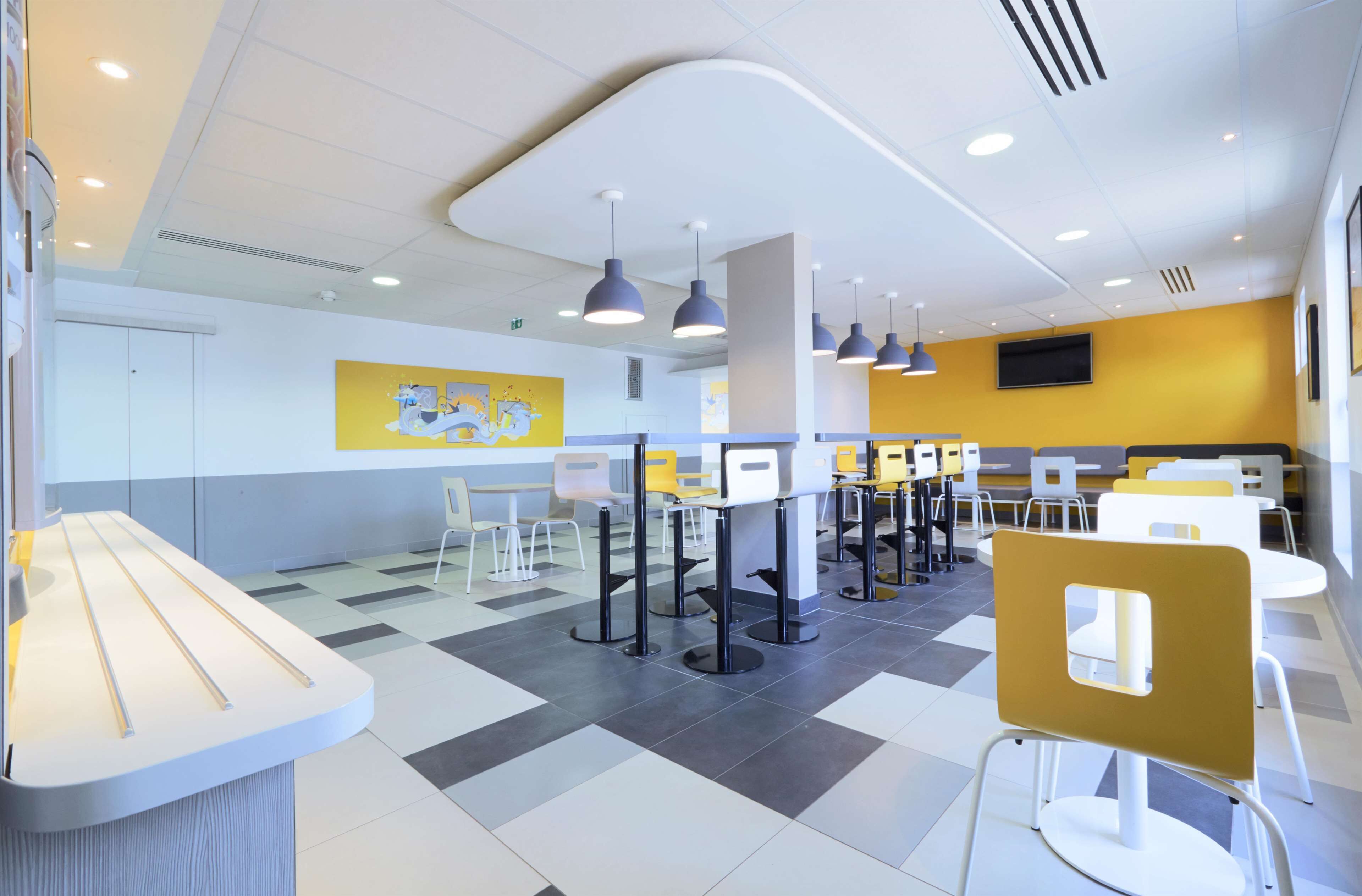 A modern cafeteria with multiple tables and chairs, a counter with a menu display, and a yellow wall with a mural.