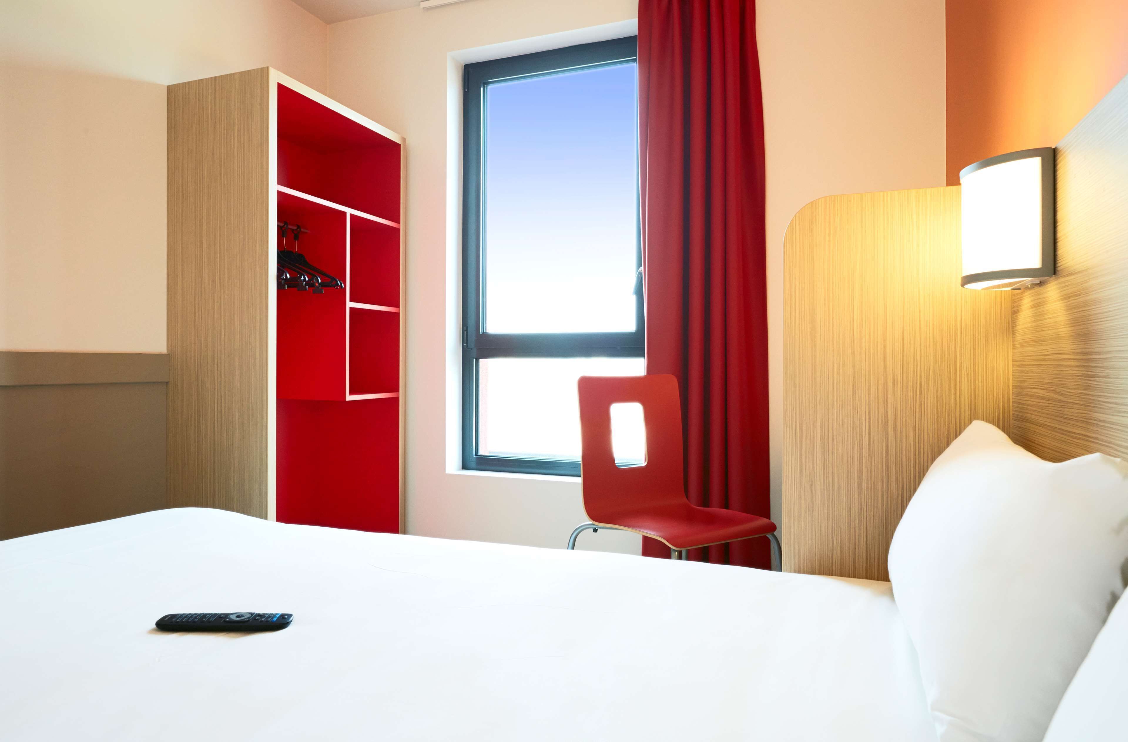 Una habitación de hotel con una cama, una silla roja, un armario y una ventana con cortinas.