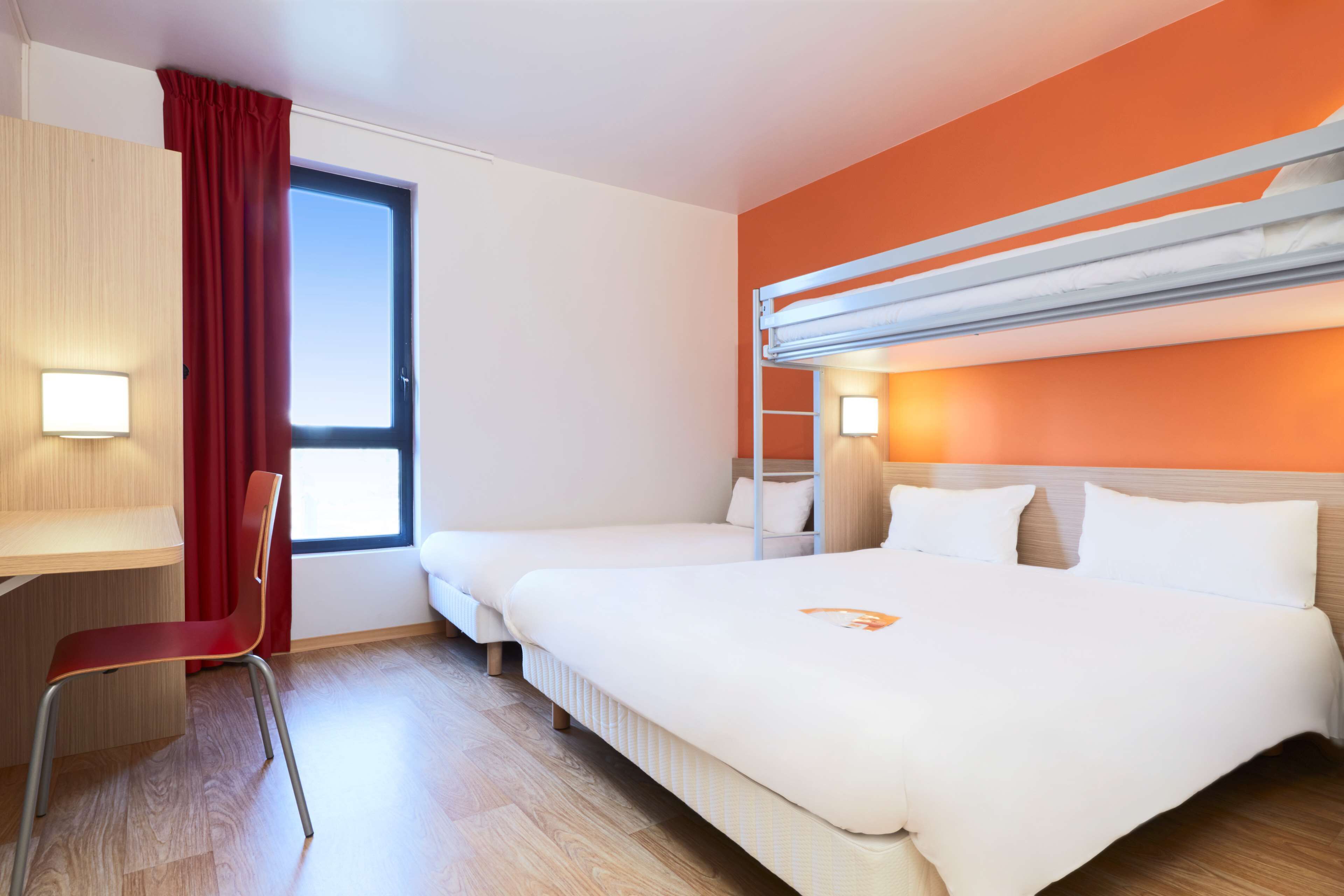 Quarto de hotel com duas camas, uma escrivaninha, uma cadeira e uma janela com cortinas.