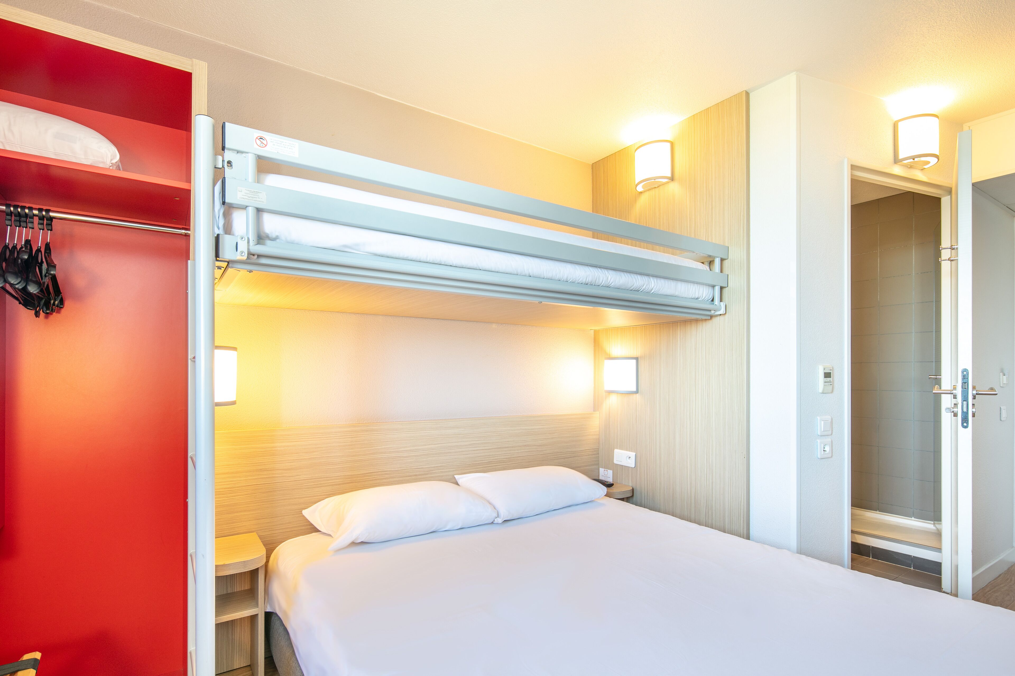 A hotel room with a bunk bed, a bedside table, a nightstand, and a bathroom door.