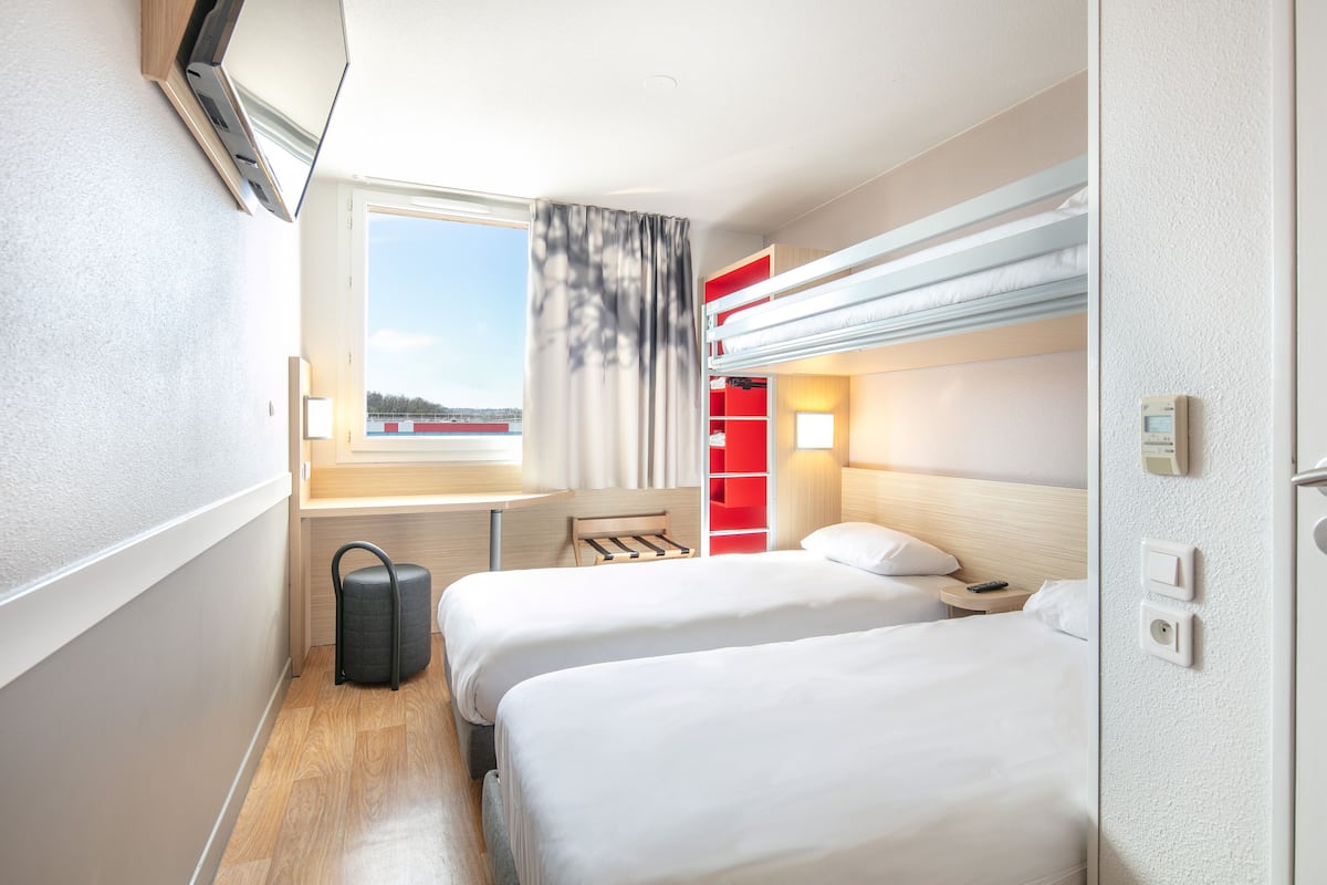 A hotel room with two beds, a desk, a chair, a suitcase, a window with curtains, a red storage unit, and a thermostat.