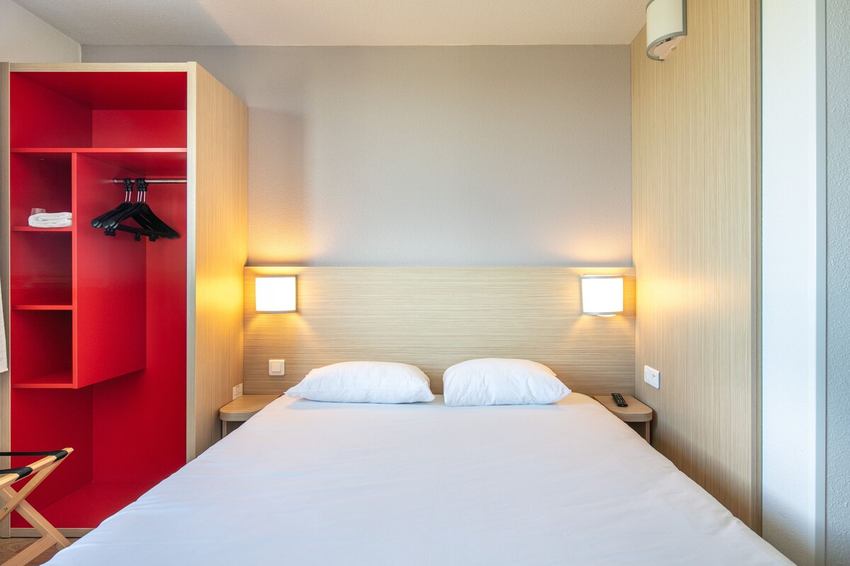 Habitació d'hotel amb un llit, dues taules de nit, un armari vermell i dues làmpades de paret.