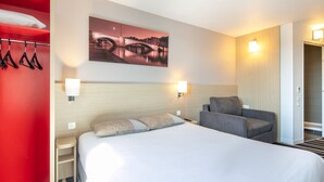 A hotel room with a large bed, a seating area with a chair, a wall-mounted painting, and two wall sconces. - B&B HOTEL Lyon Aéroport St Quentin Fallavier (Saint-Quentin-Fallavier)