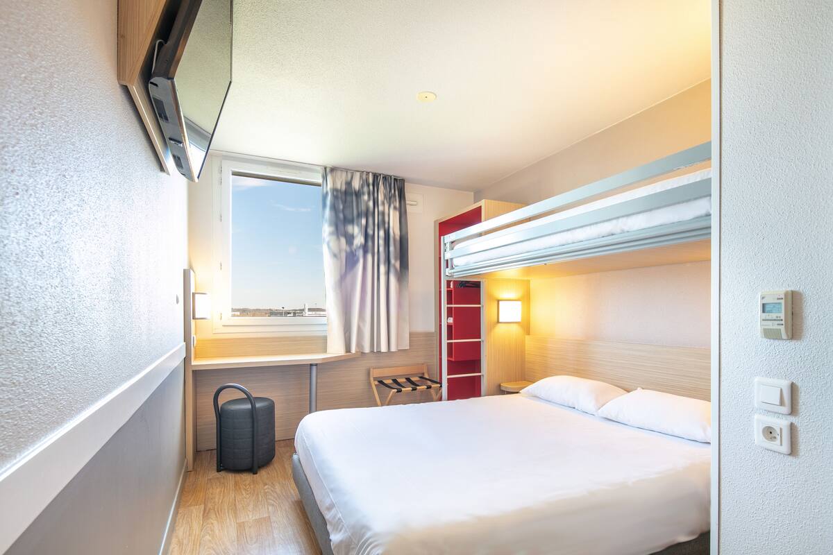 A hotel room with a bed, a desk, a chair, a suitcase, a window with curtains, a red storage unit, and a wall-mounted thermostat.