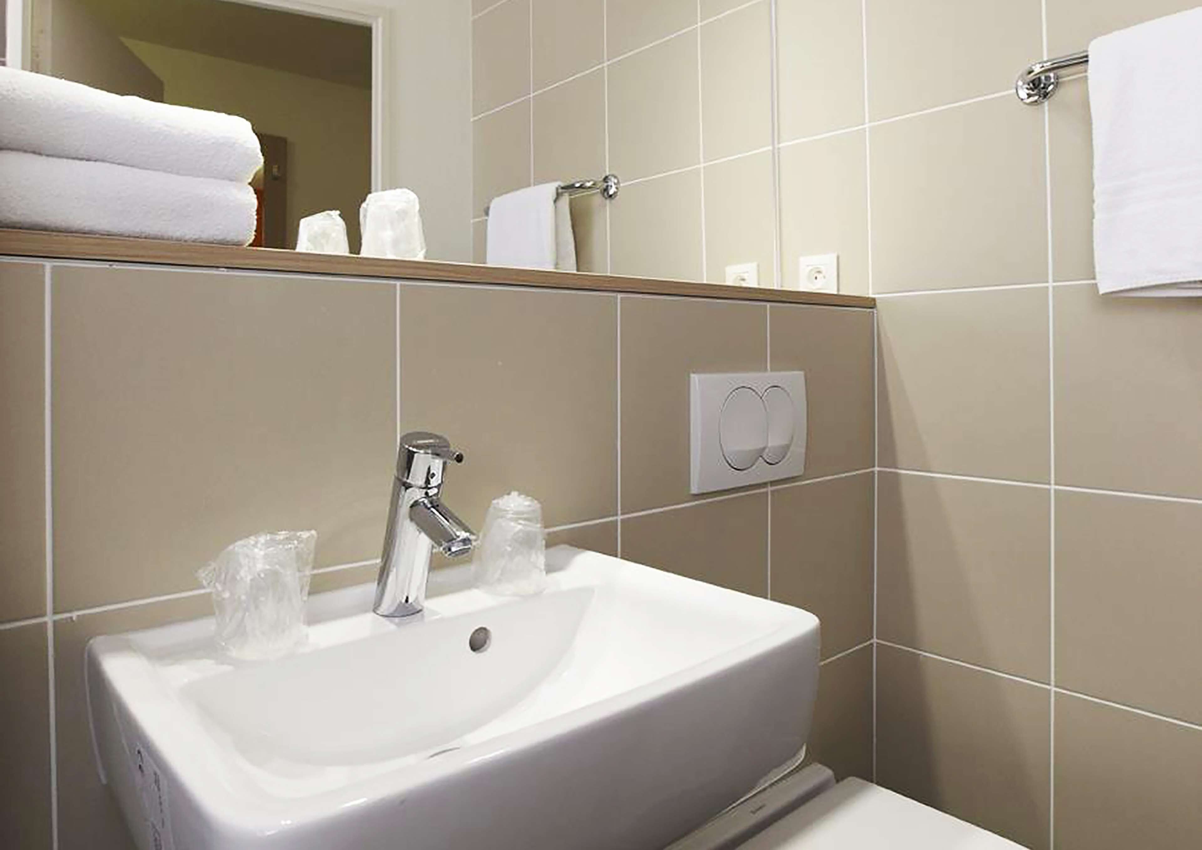 Un baño con un lavabo blanco, grifo cromado y un espejo encima. Hay dos toallas blancas apiladas en un estante y una toalla blanca colgada en un perchero.