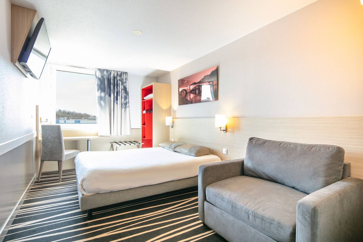 A hotel room with a large bed, a grey armchair, a desk with a chair, a red cabinet, and a wall-mounted TV.