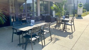 An outdoor seating area with tables and chairs, a signboard for B&B Hotels, and a building in the background. - B&B HOTEL Lyon Aéroport St Quentin Fallavier (Saint-Quentin-Fallavier)