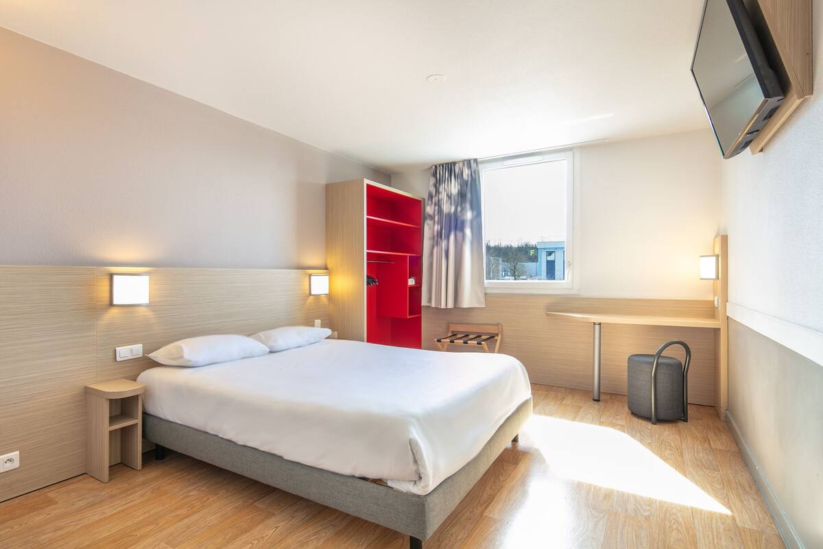 A modern hotel room with a bed, a red wardrobe, a small table, a chair, a TV mounted on the wall, and a window with a view.