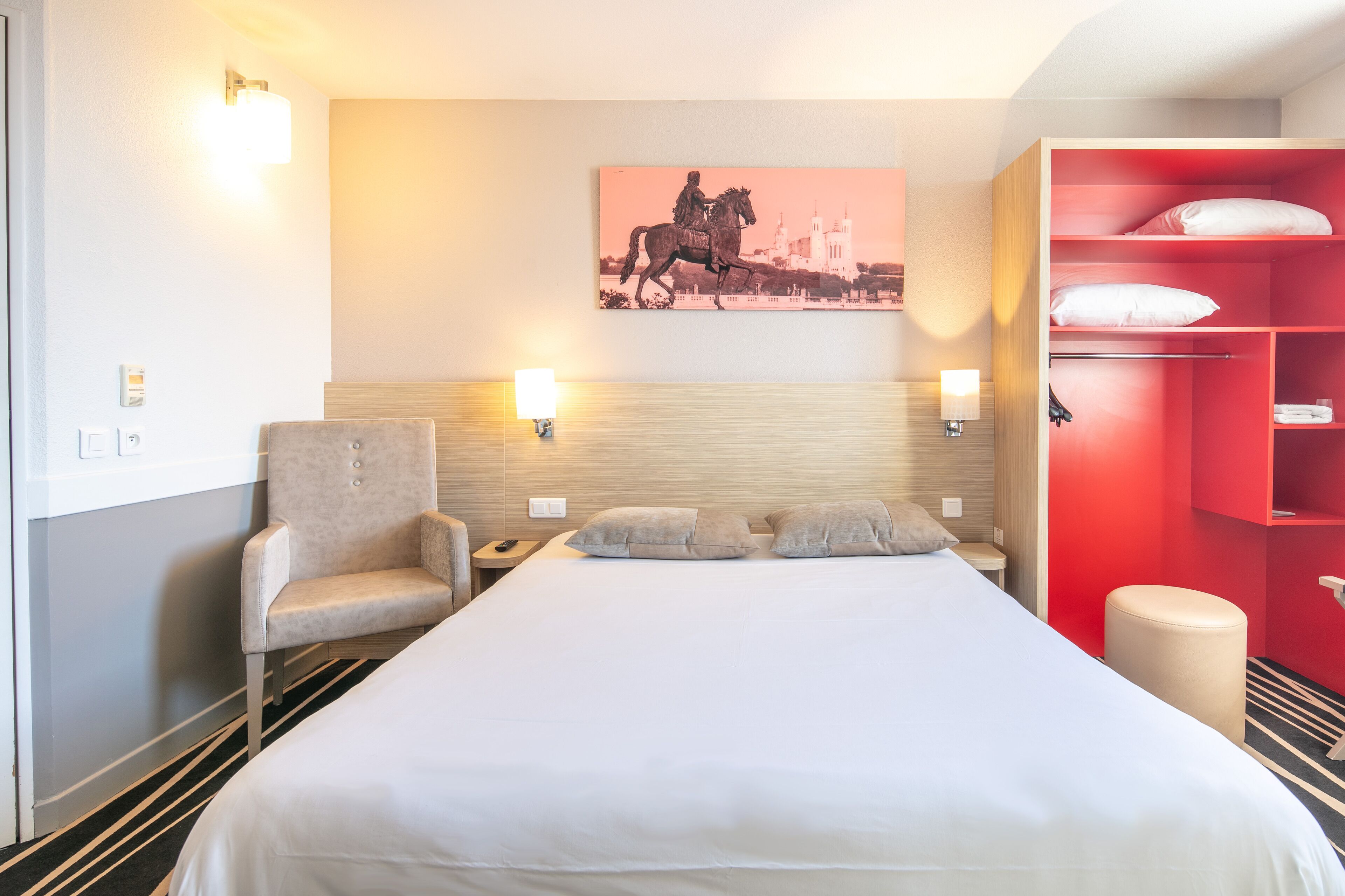A hotel room with a large bed, a chair, a red wardrobe, and a wall-mounted artwork.