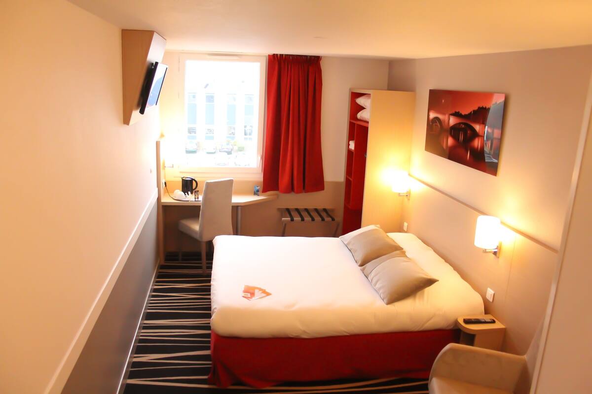 A hotel room with a bed, a desk, a chair, a window with curtains, a painting on the wall, and a lamp.