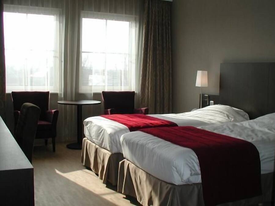 Ein Hotelzimmer mit zwei Betten, einem Schreibtisch, einem Stuhl, einem kleinen Tisch und einem Fenster mit Vorhängen.