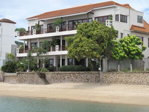 A multi-story building with balconies, a stone wall, and a beach in the foreground.