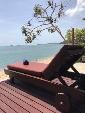 A wooden deck with a lounge chair and a towel, overlooking a beach and the ocean.