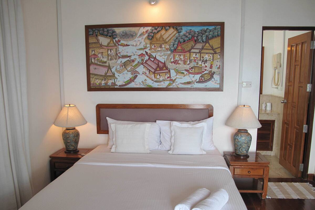 A neatly made bed with white linens and two rolled towels, a wooden headboard, two bedside tables with lamps, and a large painting of a traditional scene on the wall.